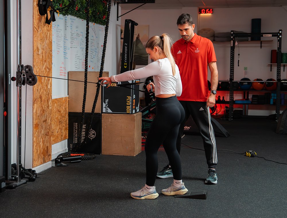 apex benifaio historia Entrenador personal supervisando técnica de ejercicio de remo con polea de una mujer en el gimnasio.