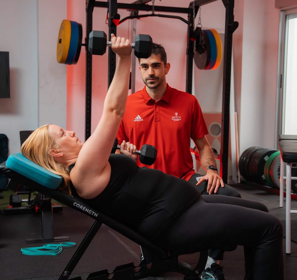 apex entrena benifaio Entrenador personal supervisando técnica de press de banca con mancuernas de una mujer en el gimnasio.