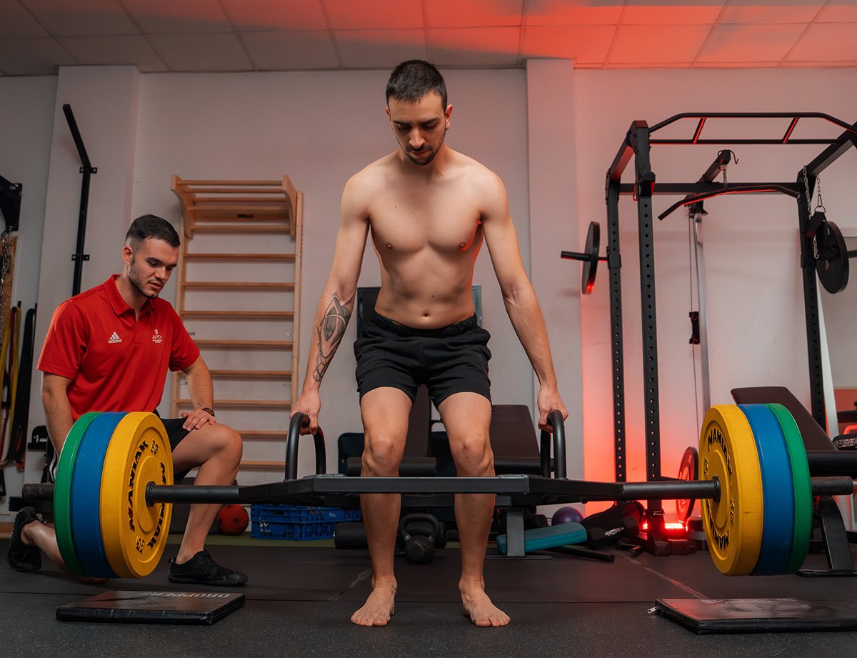 Deportista realizando ejercicio de fuerza con barra trap bar bajo supervisión de un instructor.