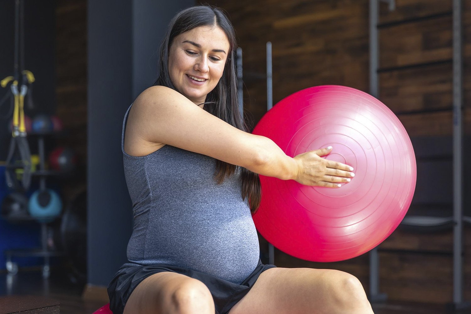 Mujer embarazada realizando ejercicio de movilidad con pelota de yoga en el gimnasio.